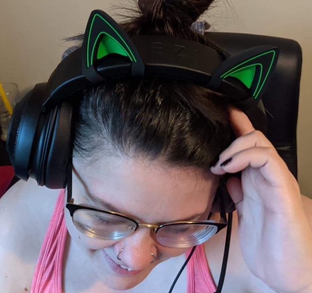 PansiePants, a white female-presenting person with glasses, looks down while touching her headset, which has green cat ears.